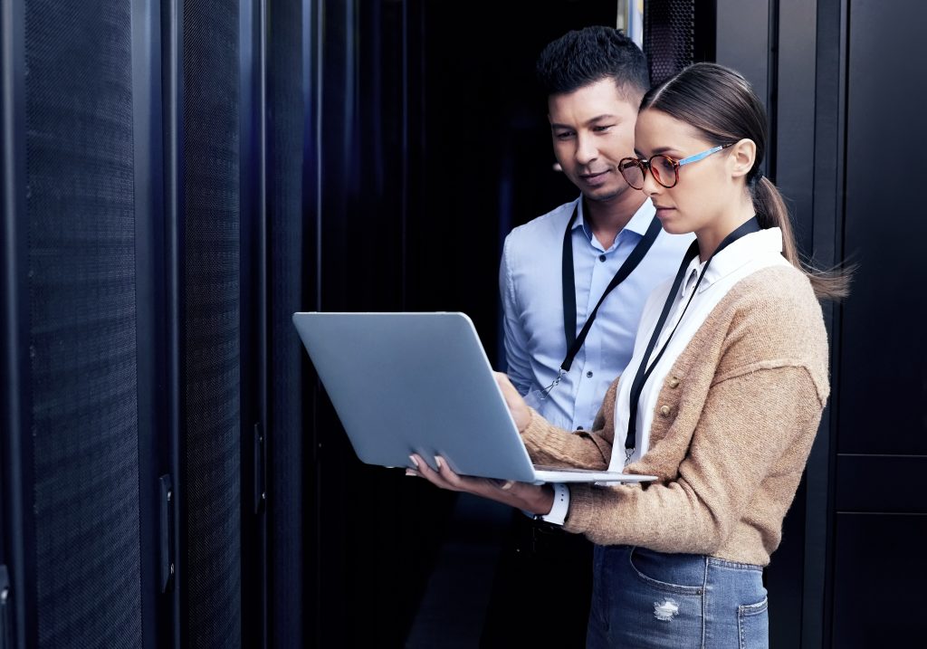 A man and woman in a server room examine a laptop in the woman's hands.